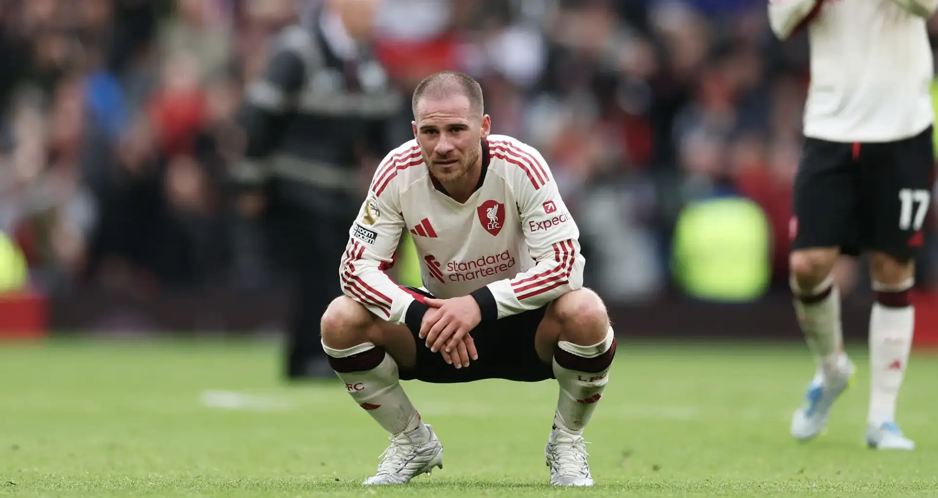 Liverpool player in a white away kit crouching on the grass during a match, with a blurred crowd in the background