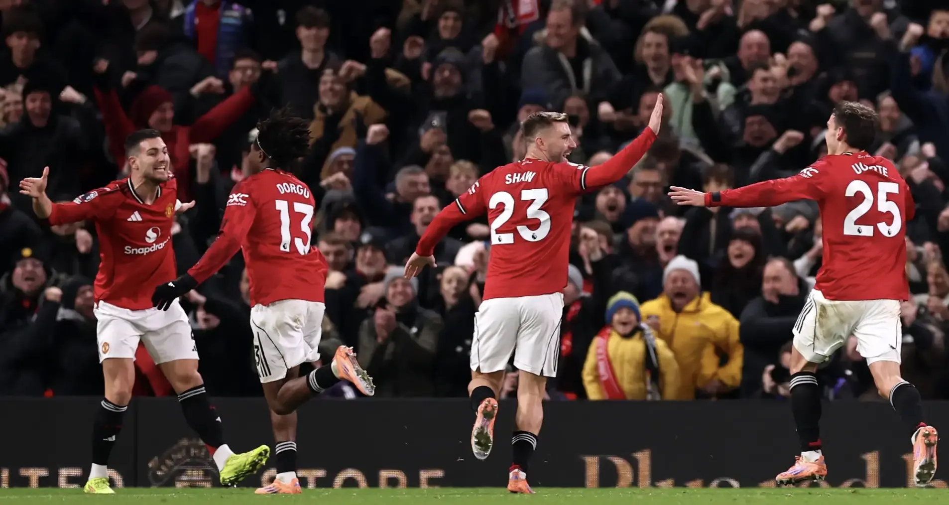 Manchester United players in red kits celebrate a goal on the pitch as fans cheer in the background.