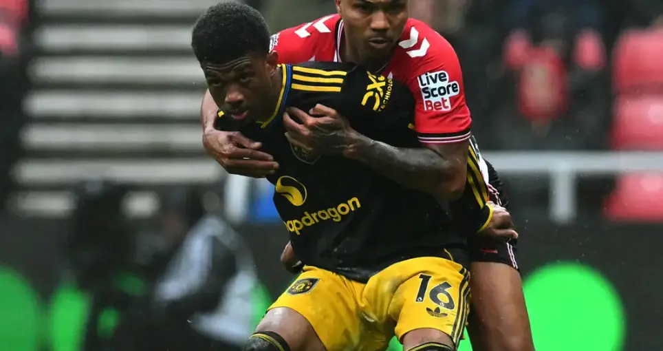 Two soccer players struggle for the ball during a match, one in a black-and-yellow kit with number 18 being held by a red-and-white kit player behind him.