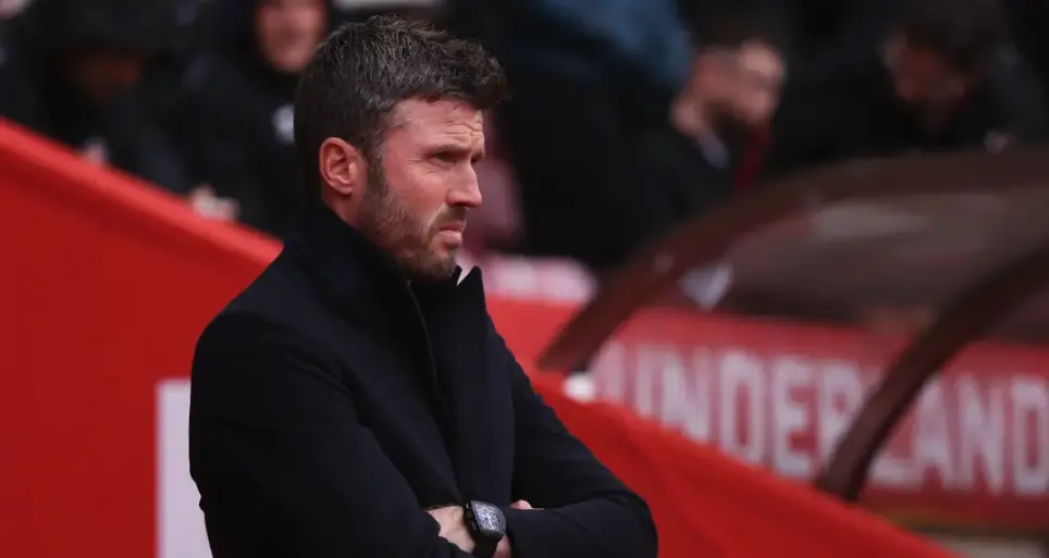 Man with short dark hair and stubble in a black coat, arms crossed, watching a game from the sideline.