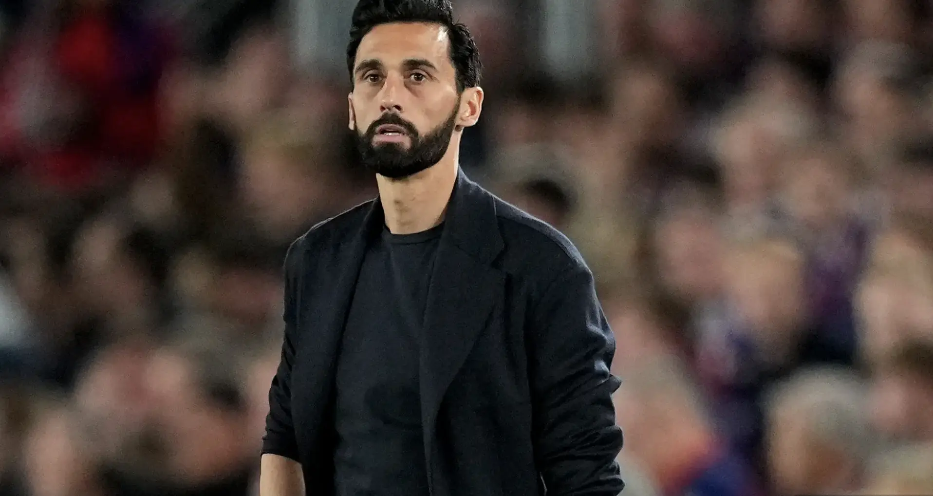 Man with dark hair and beard in a black jacket standing in a blurred crowd at a sports event
