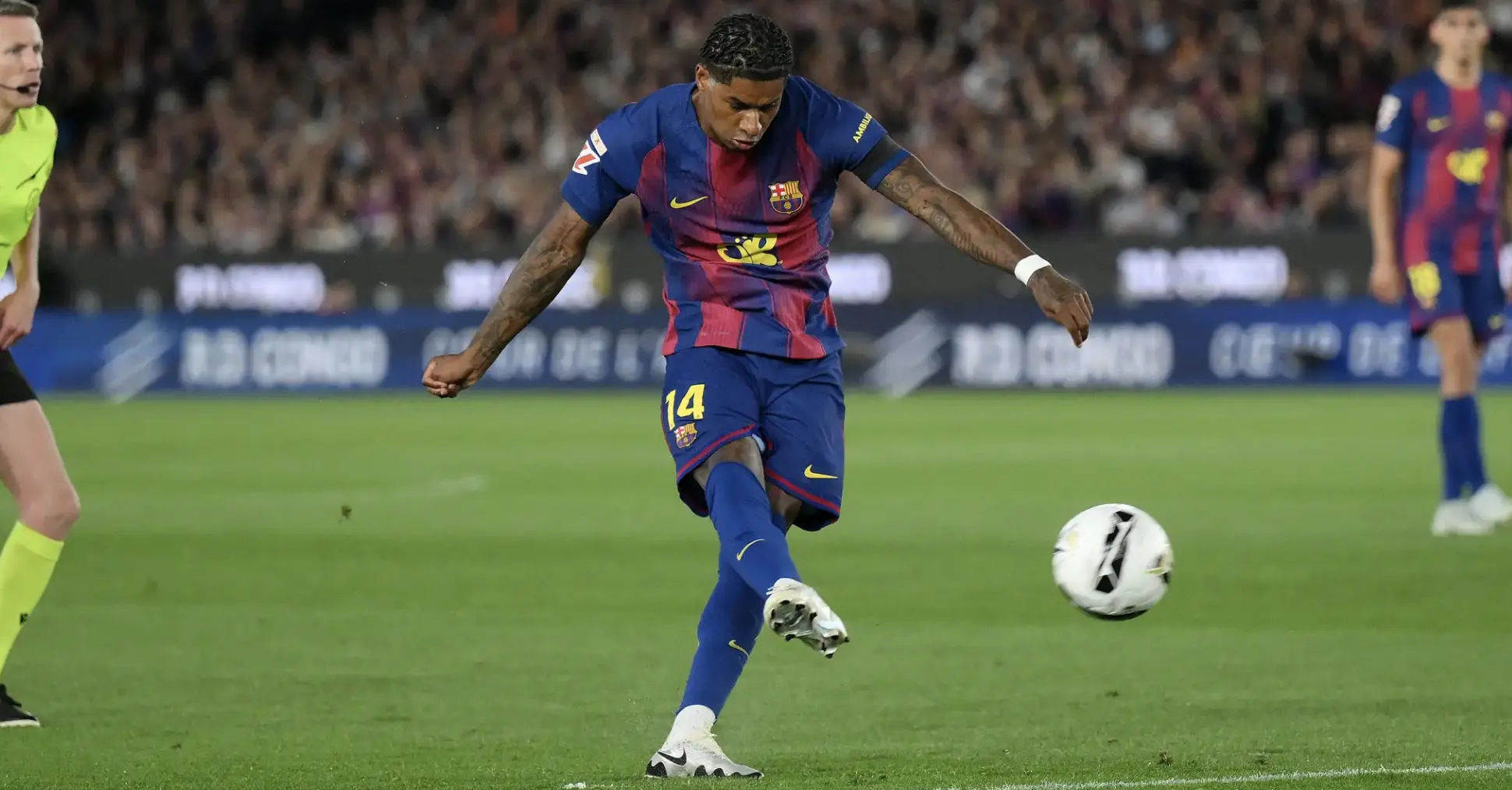 Barça footballer in blue and red kit (number 14) kicks a soccer ball during a match as fans watch in the stands behind him.