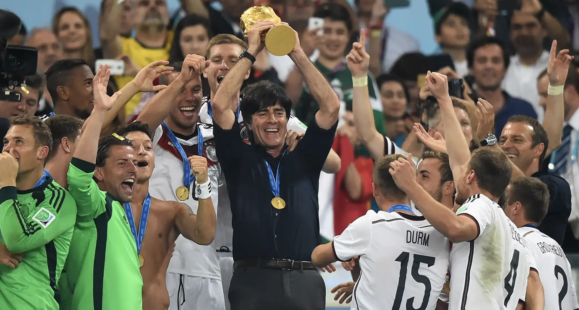 Athletes celebrate win as a coach lifts a gold trophy overhead amid cheering teammates and fans with medals around their necks.