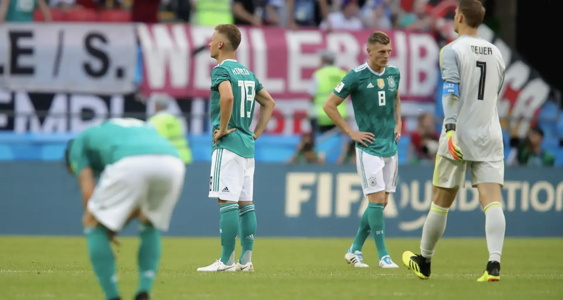 German national team players in teal jerseys stand with hands on hips on the soccer pitch, goalkeeper in gray on the right.
