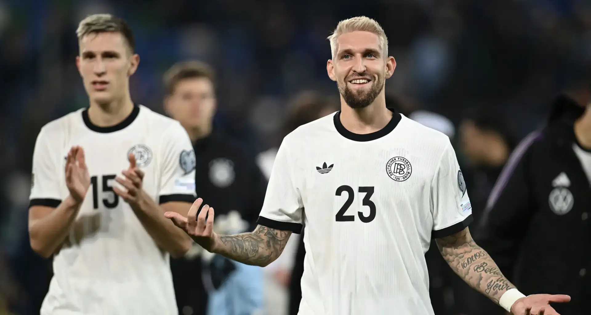 Soccer players celebrate on the pitch; foreground player in white jersey #23 with tattooed forearms smiles at camera.