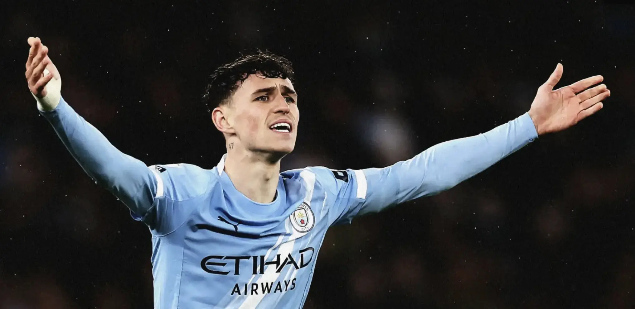 Manchester City footballer celebrating with arms outstretched on the pitch, in a light blue kit.