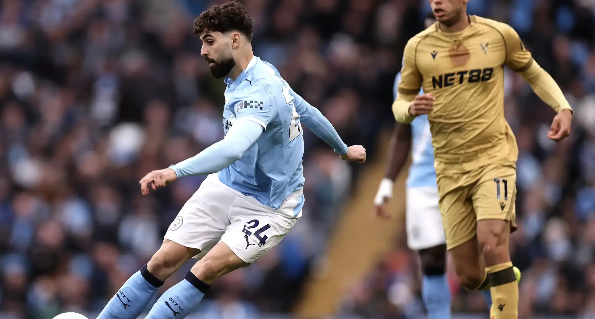 A soccer player in a light blue Manchester City kit kicks the ball while an opposing player in gold pursues from behind on a crowded pitch.