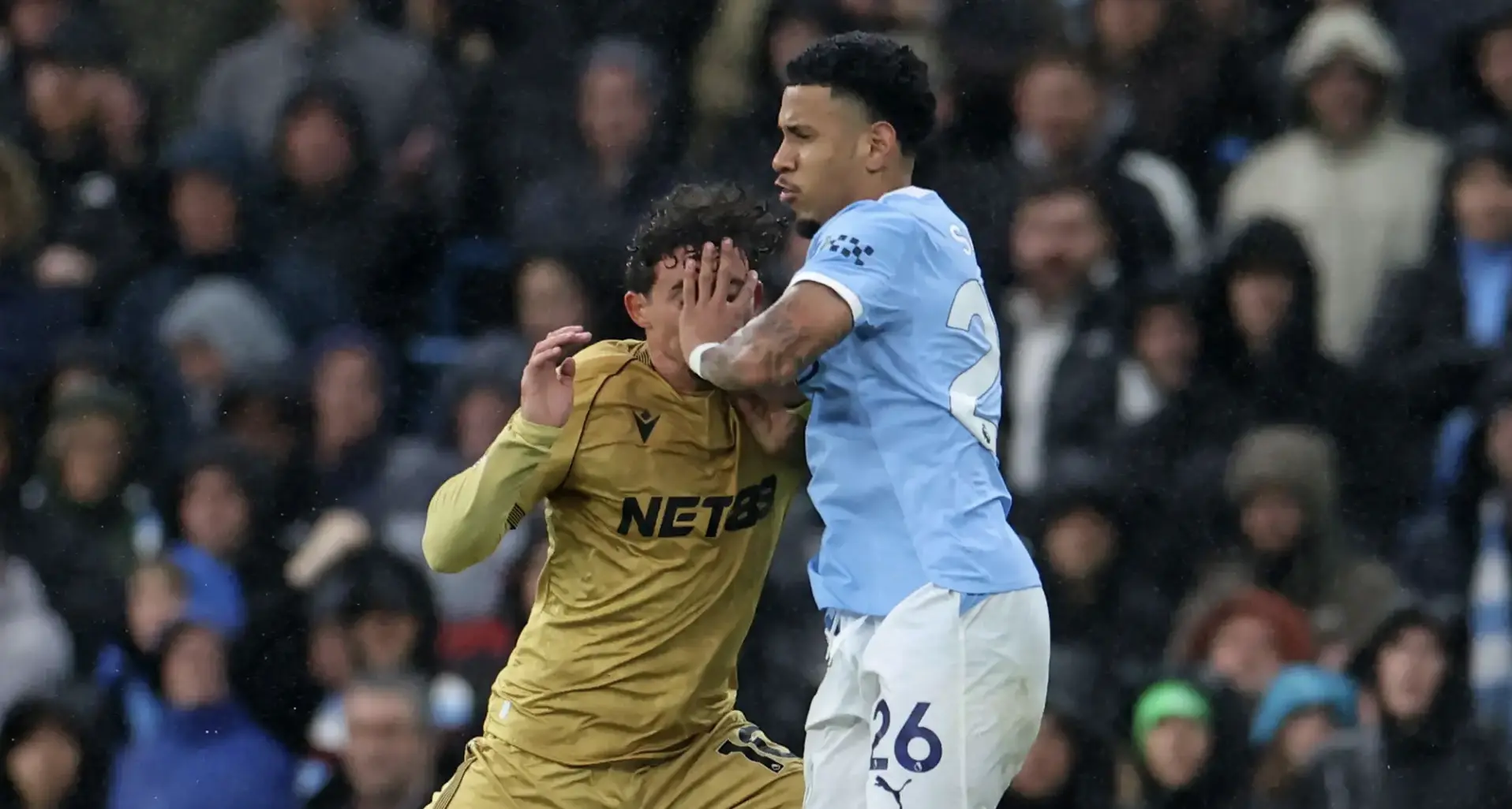 Two soccer players collide mid-action during a match, one in light blue and the other in gold, with a blurred crowd behind them.
