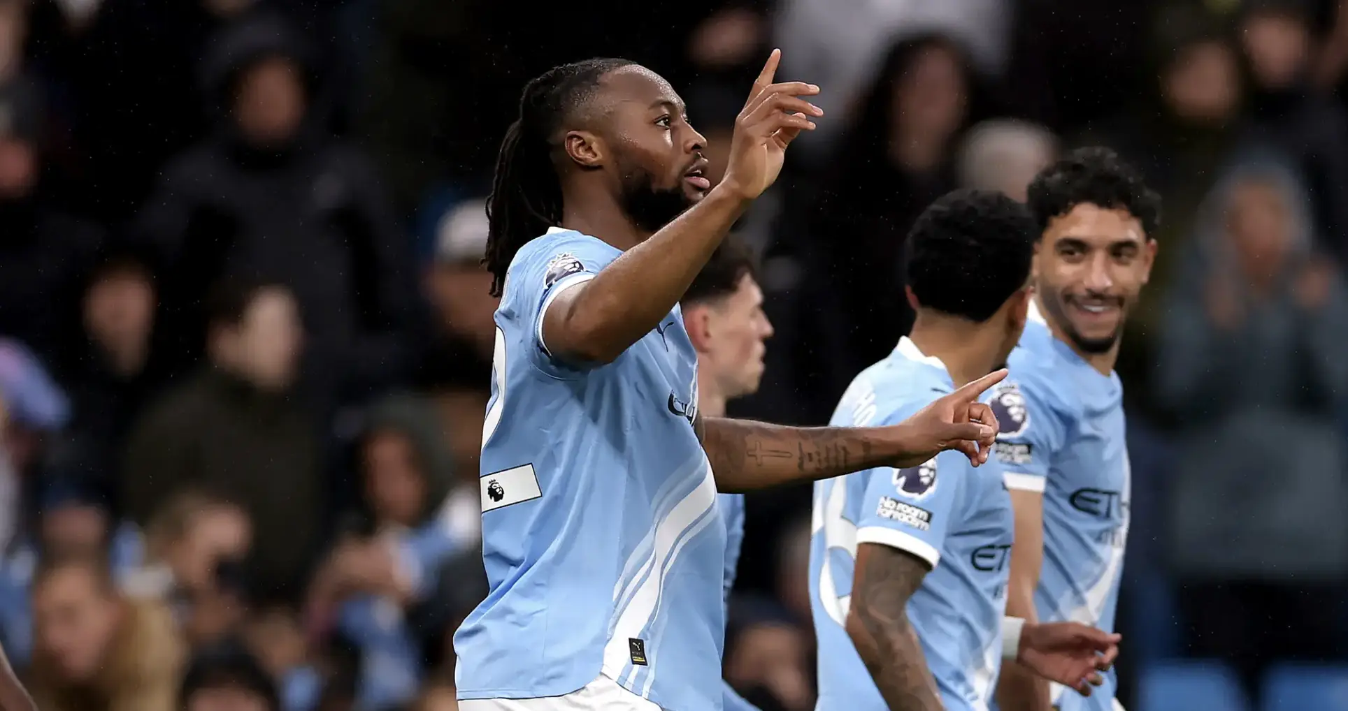 Soccer players in light blue jerseys celebrate a goal, pointing and smiling on the pitch as fans cheer in the background.