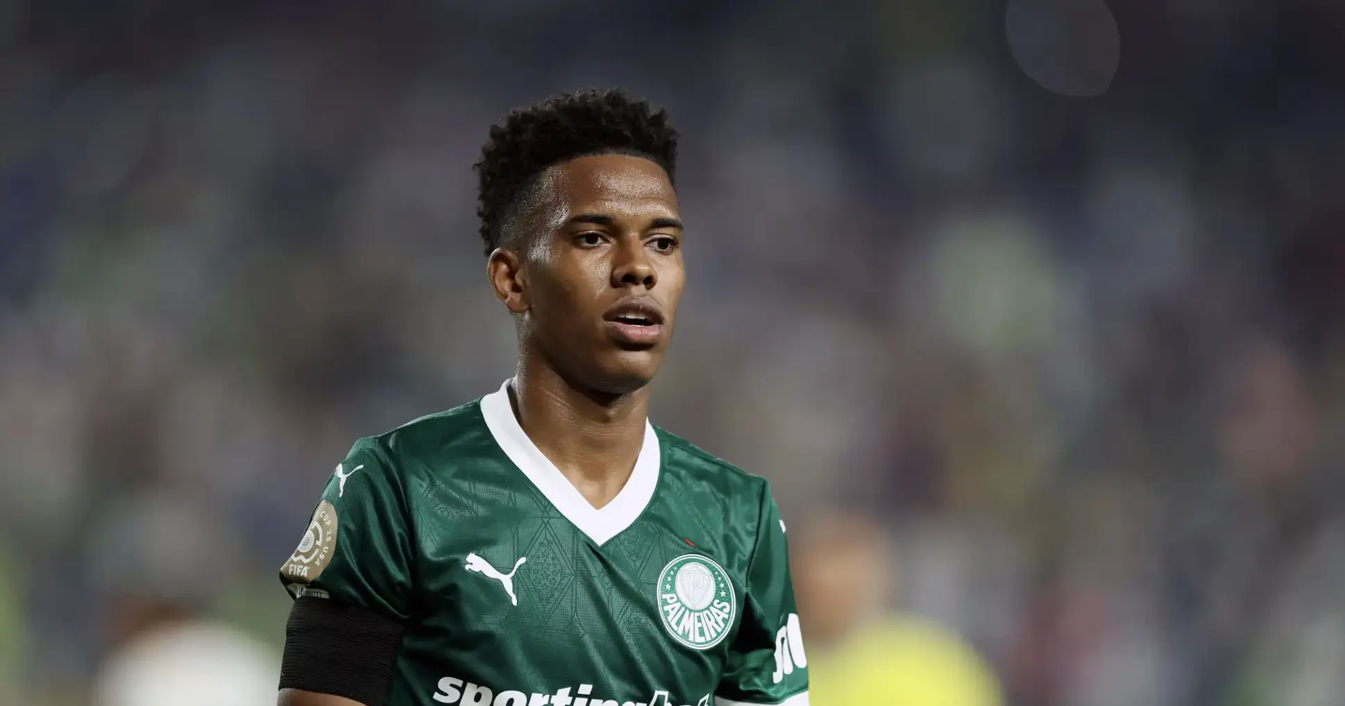 Soccer player in a green Palmeiras jersey on the field, looking to the side with a blurred crowd behind him