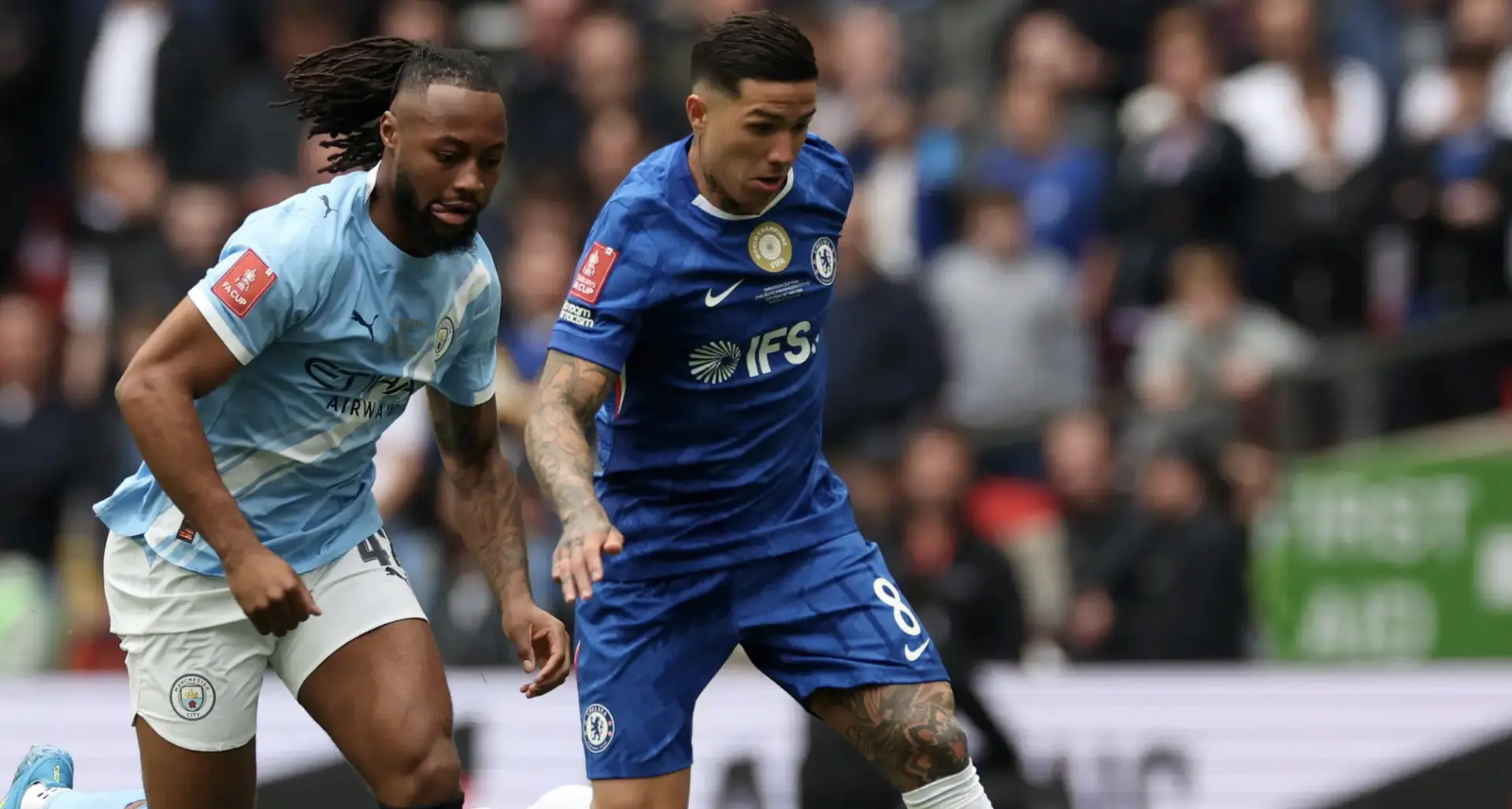 Two soccer players in blue kits chase the ball during a crowded match, with Manchester City and Chelsea in action.