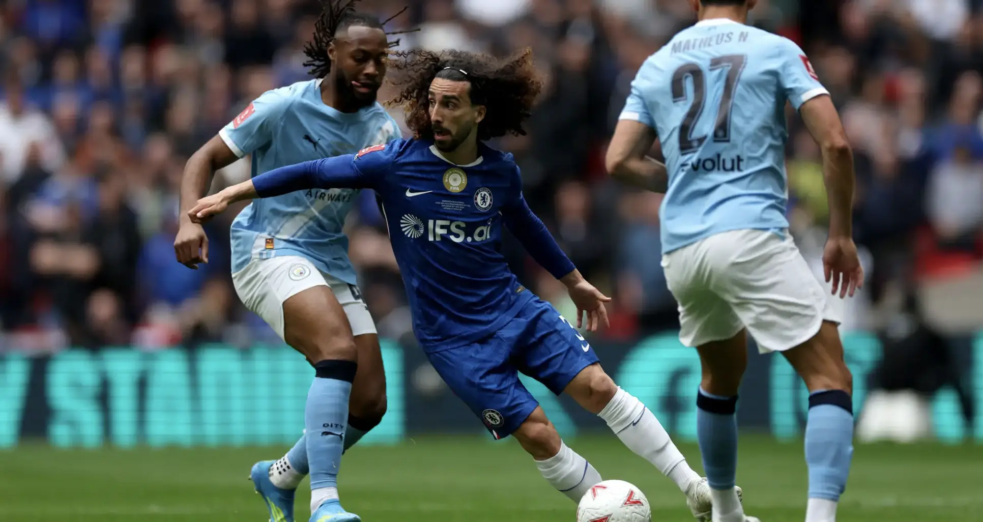 Chelsea player in blue controls the ball while two opponents in light blue chase him on a crowded pitch.