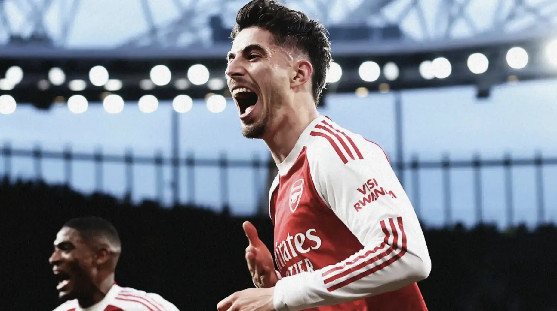 Arsenal player in red-and-white kit celebrating with mouth open, stadium lights behind him.