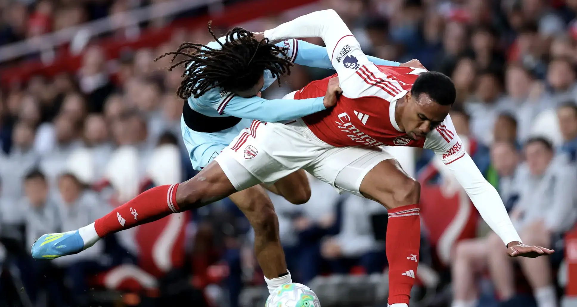 Two soccer players duel for the ball in a crowded stadium, one in Arsenal red-and-white, the other in light blue goalkeeper kit.