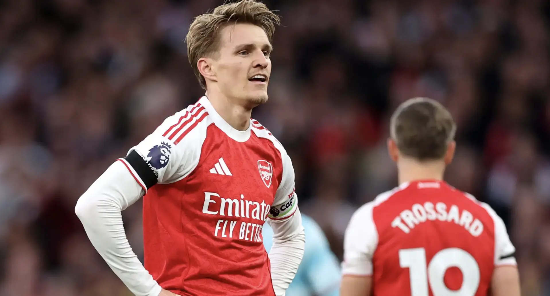 Arsenal players in red and white kits on the pitch; one faces the camera with hands on hips, the other seen from behind with 'TROSSARD' on his jersey.