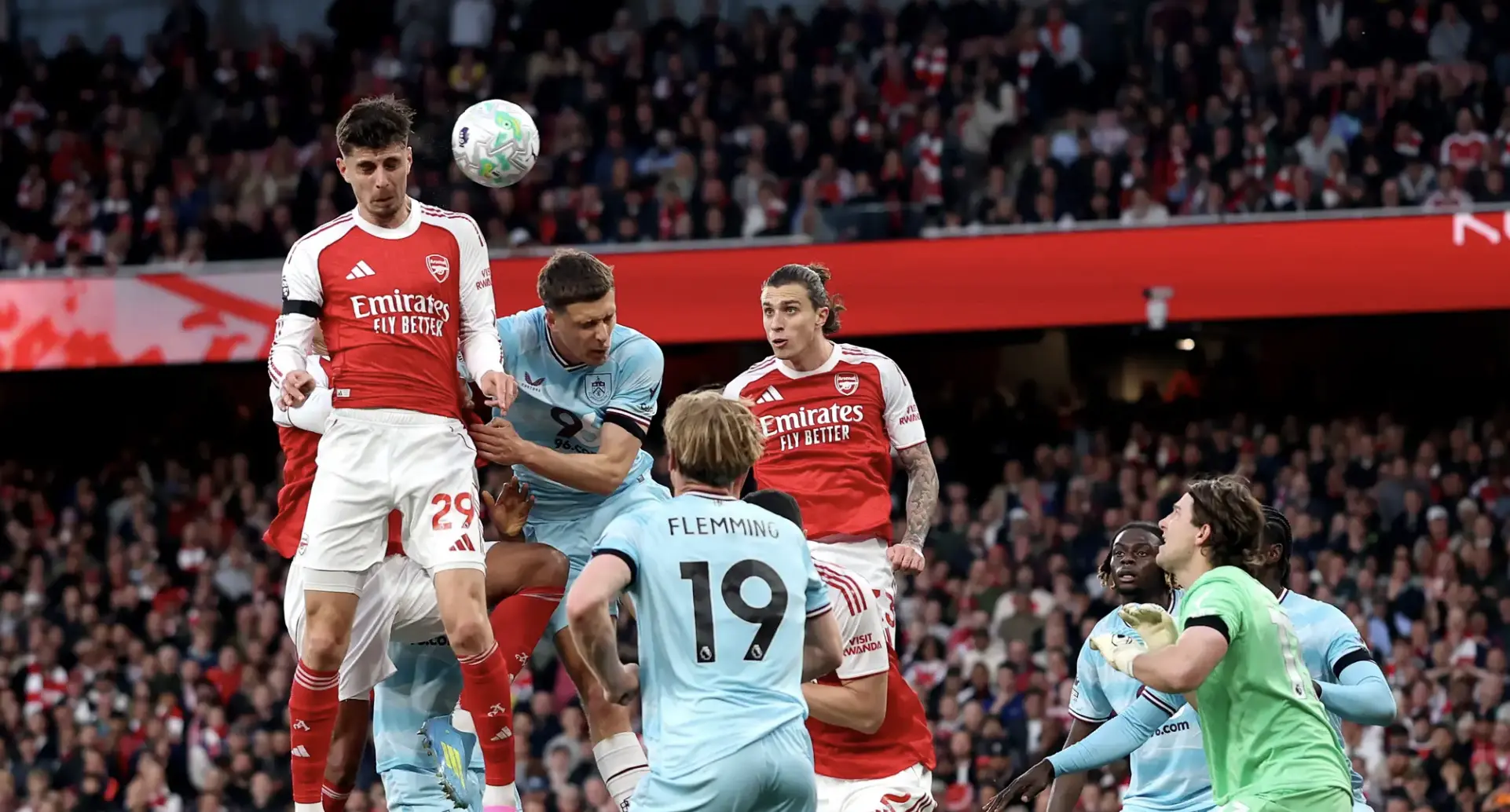 Arsenal players in red fight for a header against light-blue defenders with the ball in the air amid a full stadium crowd.