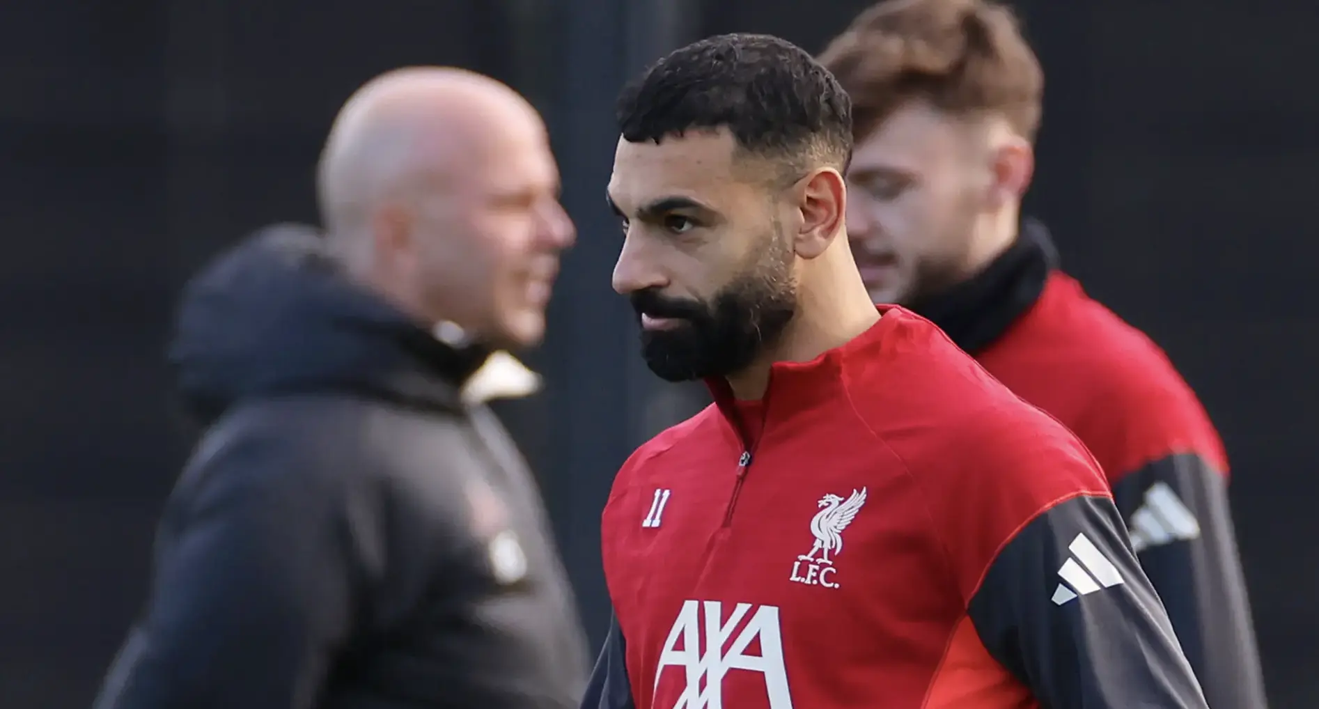 Close-up of a Liverpool FC player in red training gear during practice, with teammates/coaches blurred in the background.