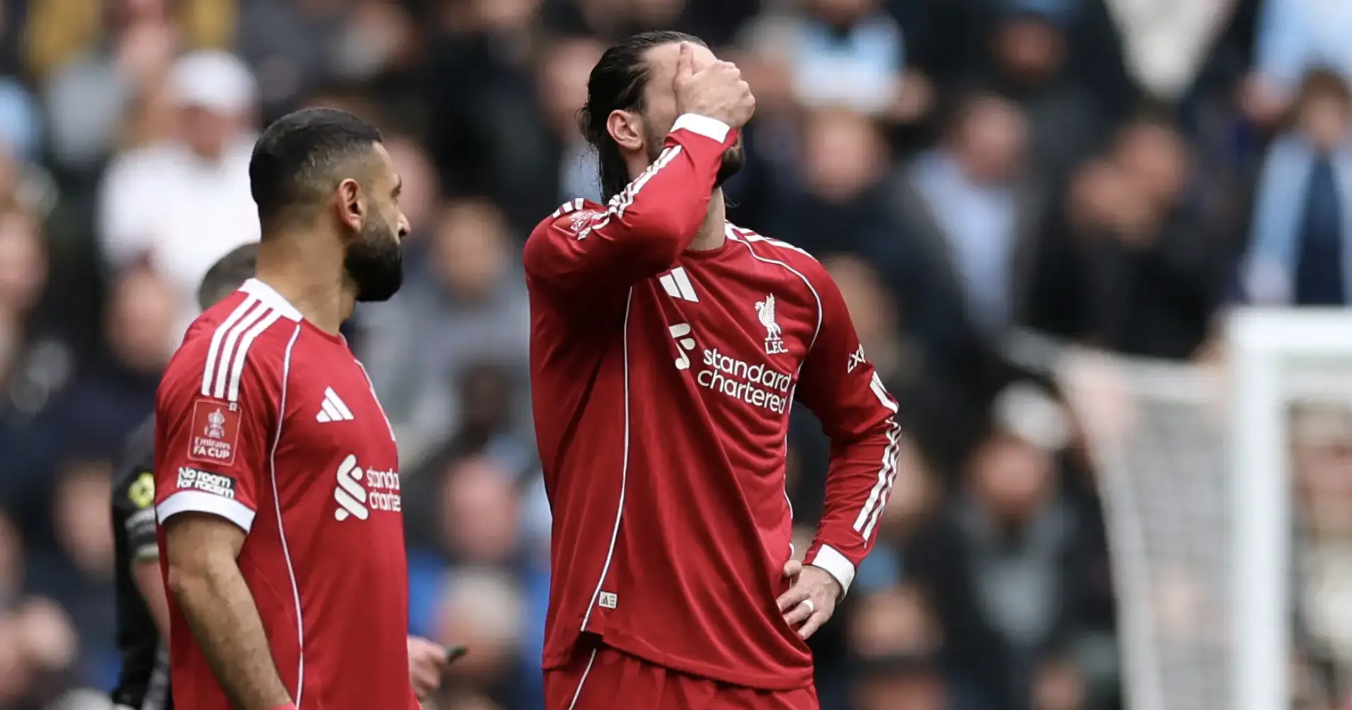 Liverpool players in red jerseys on the pitch; one covers his face with his hand while the other looks on, with a blurred crowd in the background.