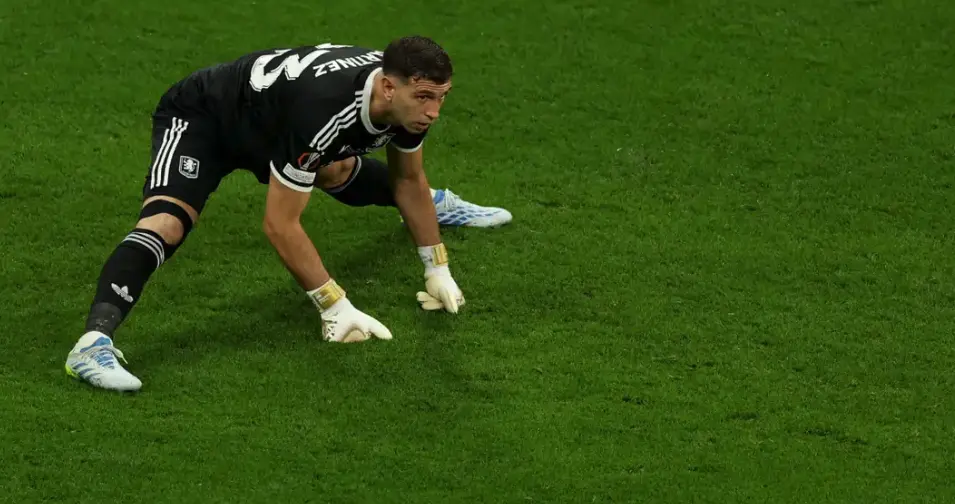 Soccer goalkeeper in a black kit crouching on a green pitch, wearing gloves and ready to defend the goal.