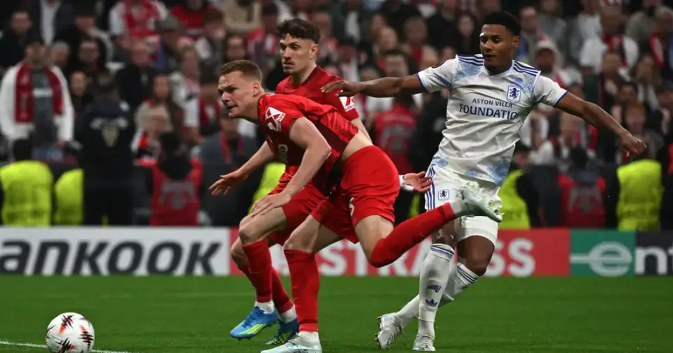 Two red-clad players contest the ball with a white Aston Villa player during a crowded stadium match moment on a green pitch.