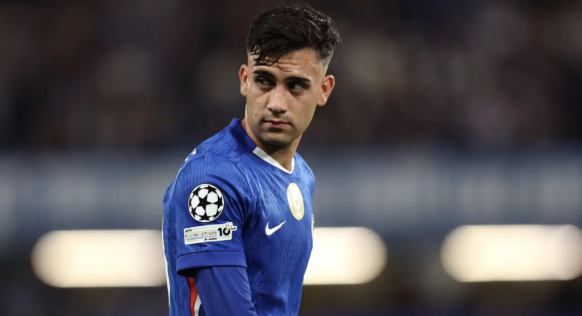 Male footballer in a blue jersey looking to his left on the pitch, Champions League badge visible on sleeve.