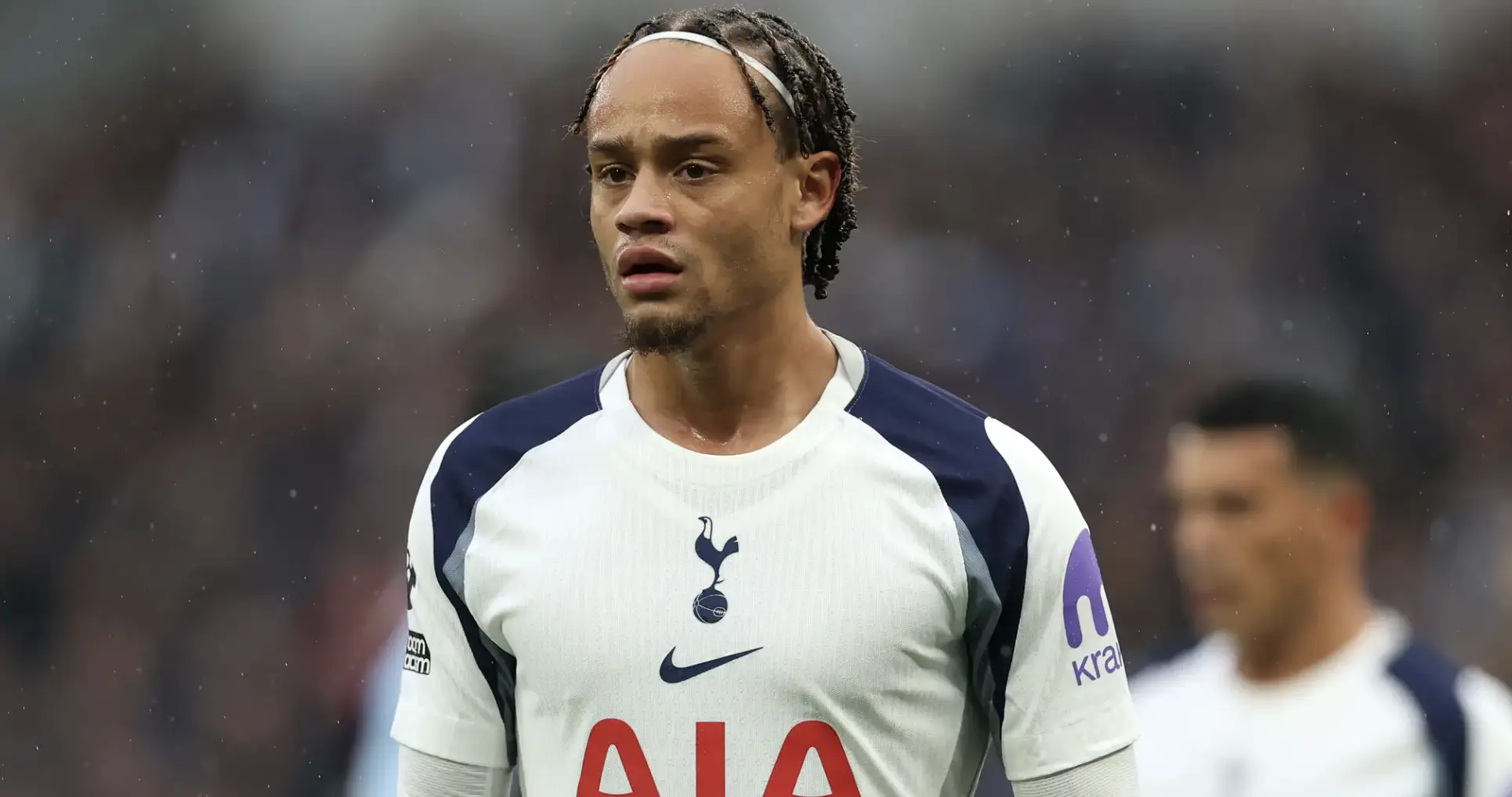 Tottenham Hotspur footballer on the pitch in a white kit with navy accents, looking focused as it rains in the background.
