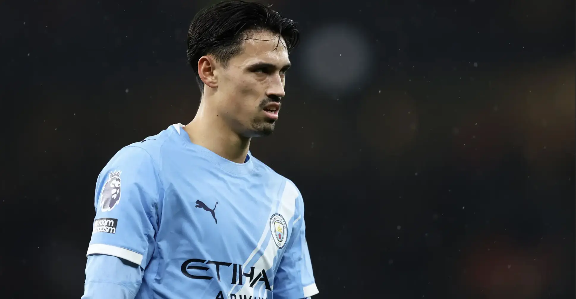 Manchester City footballer in light blue kit on the pitch, focused during a match in the rain.