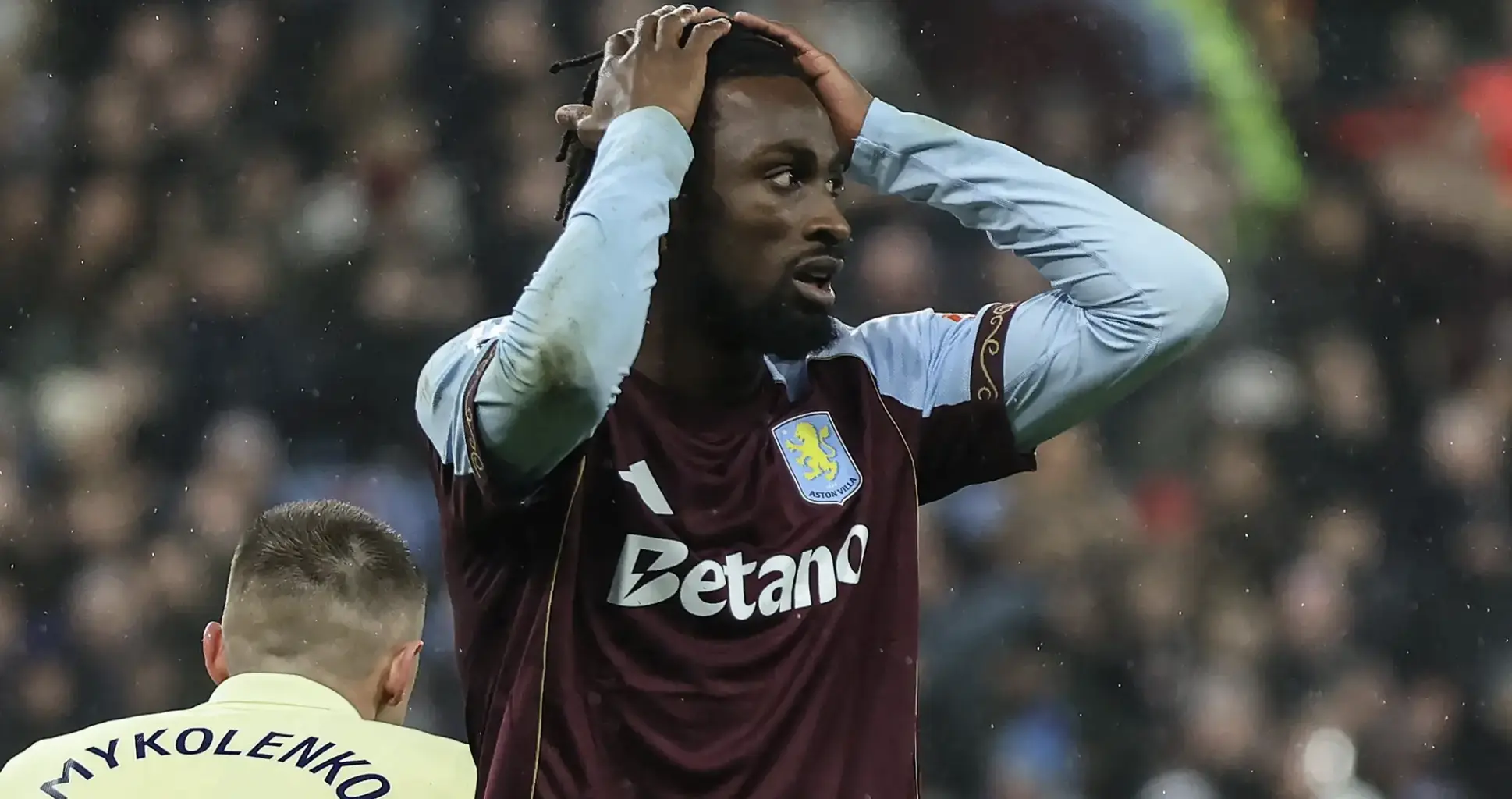 Aston Villa footballer in maroon and light-blue kit holds his head in frustration on the pitch as rain falls.