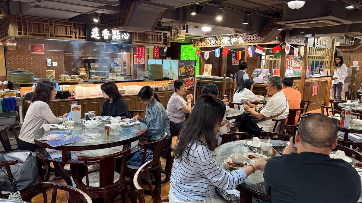 Busy Asian restaurant with round glass tables and diners eating, open kitchen visible in the background and decorative wooden panels above the counter.