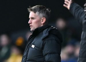 38911066835_586x4201 - 實發體育 Side profile of a man in a black puffer jacket standing on the sidelines of a sports field, looking focused.