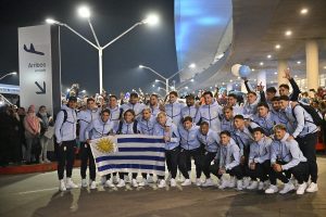 39653650461_600x400 - 實發體育 Group of athletes in light-blue jackets posing with the Uruguay flag at a night-time airport arrivals area.