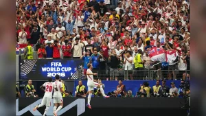 Soccer players in white celebrate on the field as a large, cheering crowd watches; FIFA World Cup 2026 sign visible in the stands.