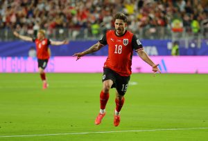 Ak0de7i29rIItXohPQUBP6H7jE9oqoW1xFlh2sRZYdo - 實發體育 Soccer player in a red and black jersey with number 18 sprinting on the field, crowd in the stands behind.