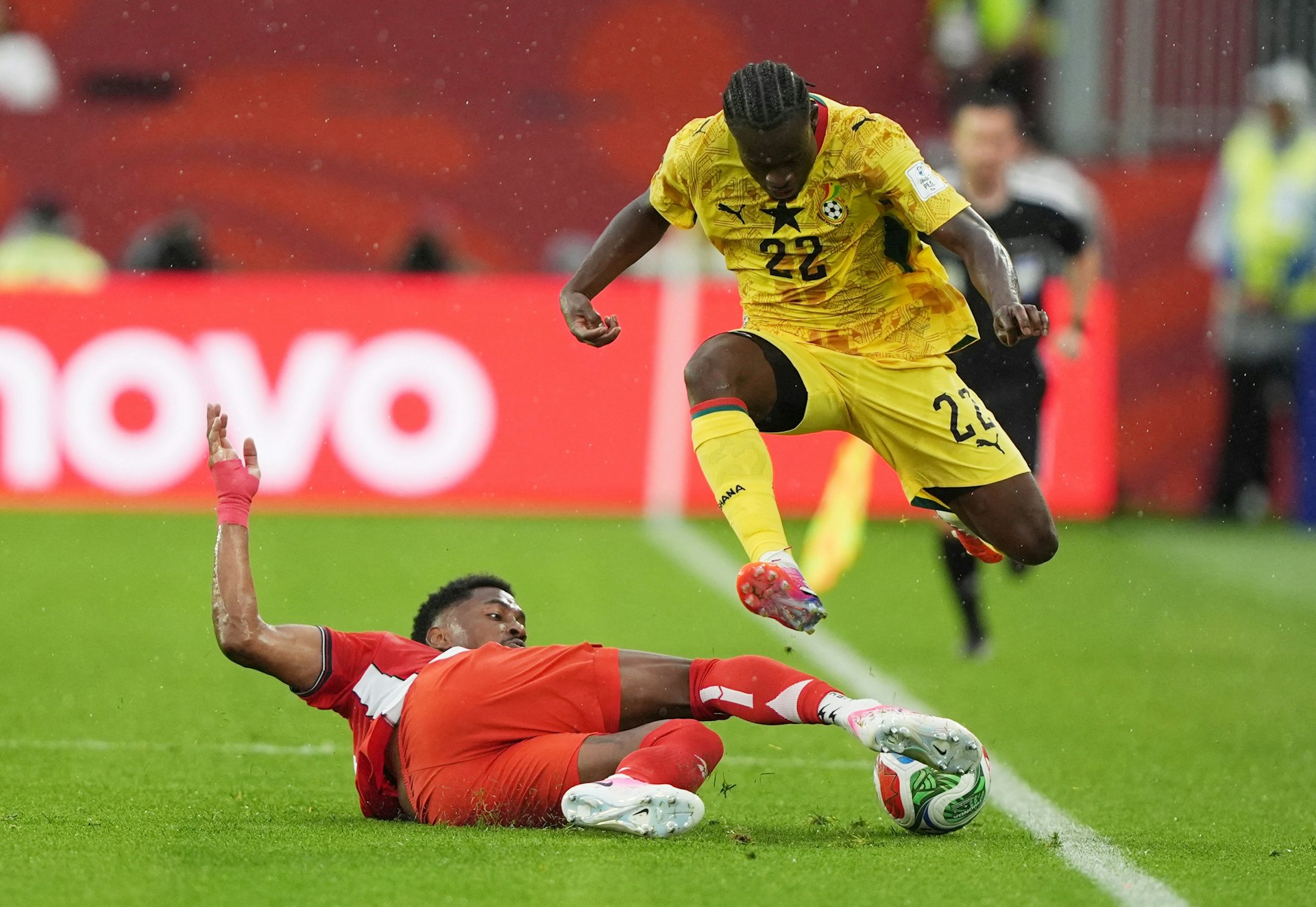 GdgYiKNkB0h7SuvrOUdEXGK-Y3Wj3ZeuXkdsD15HbA8 - 實發體育 Ghana soccer player in yellow jersey (number 22) jumps over a sliding red-team opponent during a match, ball at his feet on the green field
