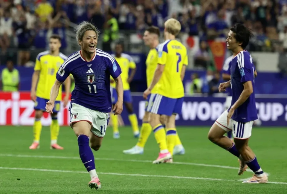 Japan national team player #11 in blue celebrates a goal on the field, with teammates and yellow-clad opponents nearby.