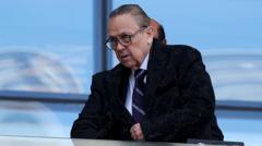 cover-7655-1780790465jpg - 實發體育 Older man with glasses in a dark suit leaning on a desk in a bright, modern office setting with blue window background.