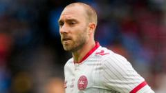 Male soccer player in a white kit with red trim on the field during a match, with a blurred crowd in the background.
