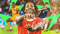 cover-7716-1781022893jpg - 實發體育 Close-up of a joyful footballer in a red jersey reaching toward the camera, with teammates celebrating on a green field in the background.