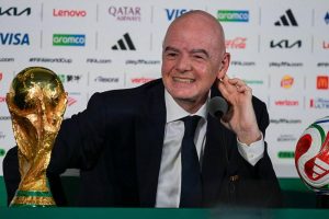 cover-7741-1781130897jpg - 實發體育 Smiling bald man in a suit sits at a press conference desk beside a golden World Cup trophy and a soccer ball on his right.