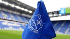 cover-7763-1781148894jpg - 實發體育 Blue flag featuring a white club crest waving in a football stadium, with green pitch and empty blue seats in the background.