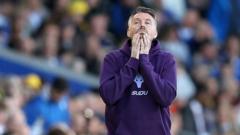 cover-7766-1781166890jpg - 實發體育 Soccer coach in a purple jacket on the sideline with hands to mouth, reacting during a match, blurred stadium and crowd in the background.