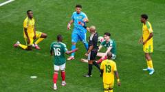Soccer players in green and yellow uniforms cluster around the ball on a green field, with a referee nearby.