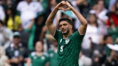Soccer player in a green jersey with number 3 forming a heart shape over his head during a match, with cheering crowd in the background.