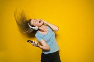 cover-7957-1781678224jpg - 實發體育 Woman wearing white over-ear headphones sings and dances with a phone in hand against a bright yellow background.