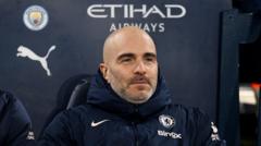 Man in a blue Manchester City jacket sitting on a dugout bench, with Etihad Airways and Puma logos visible in the background.