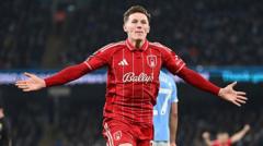 Soccer player in a red kit celebrating with arms outstretched on the field, stadium lights in the background.