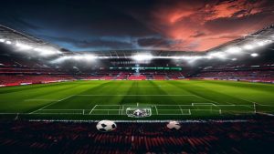 Soccer stadium at dusk with bright floodlights, red seats, and a green pitch; two balls resting near the foreground goal line.