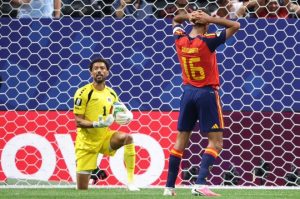 Goalkeeper in yellow kneels with the ball as a rival in red and blue stands with hands on his head near the goal net.