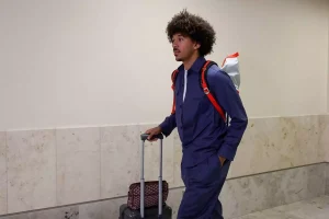 f60bf19061effed8a13346ebab28b251_US_WCup_Soccer_62944jpg1 - 實發體育 Man with curly hair in a blue outfit pulling a wheeled suitcase while wearing a red-backpack, walking through a light hallway.