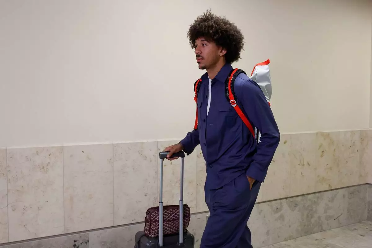 Man with curly hair in a blue outfit pulling a wheeled suitcase while wearing a red-backpack, walking through a light hallway.